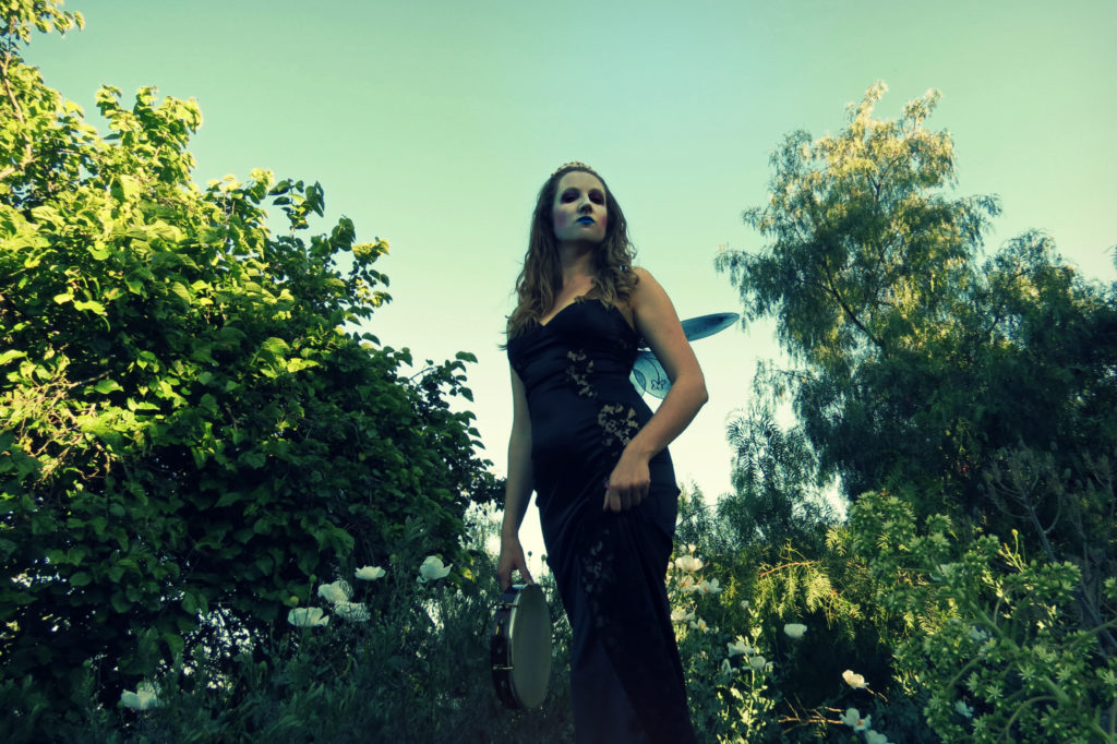 Catherine, dressed as Queen Mab, stands regally in the garden holding her pandeiro in a black form-fitting dress, tiara, and wings.