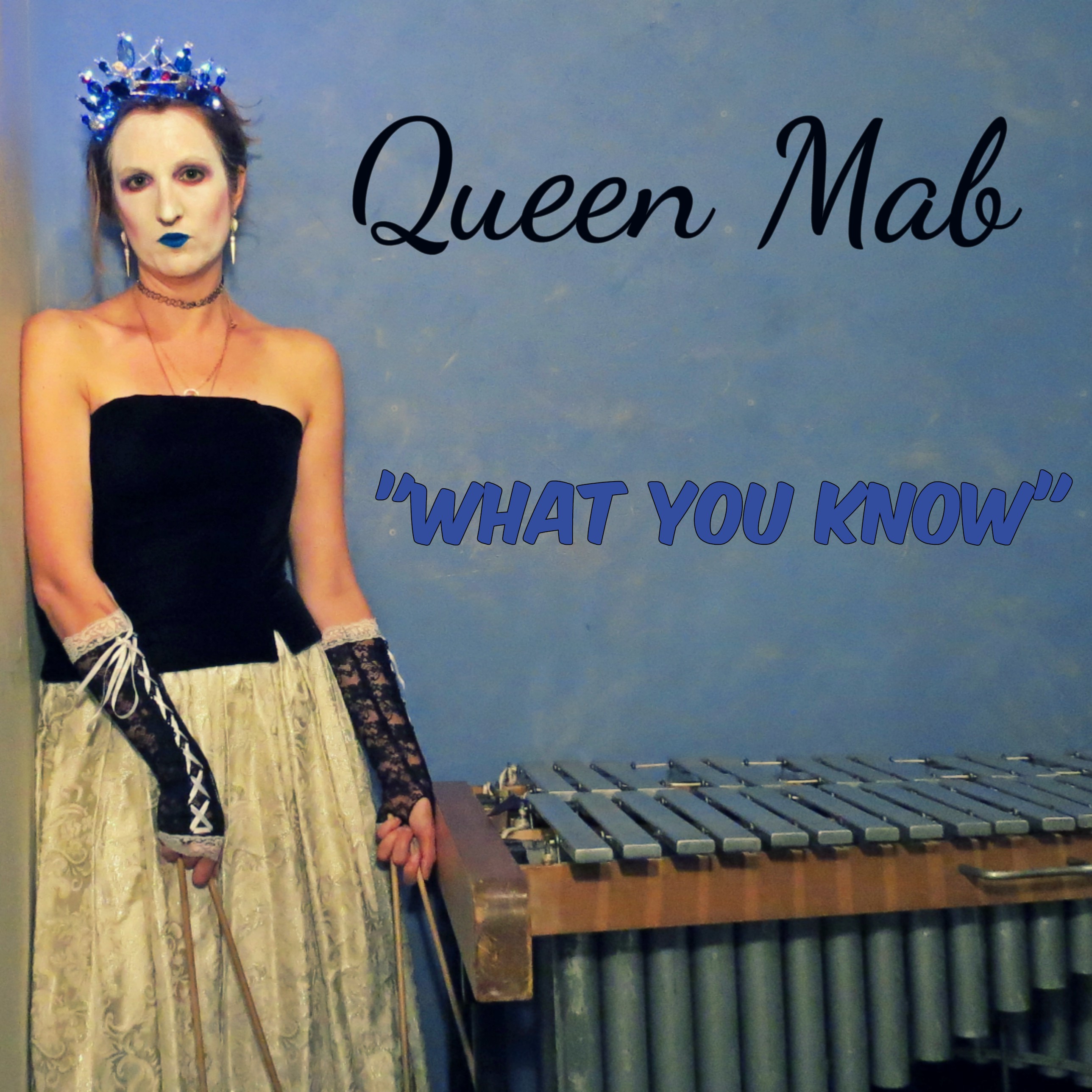 Catherine stands next to her vibraphone holding four mallets. She has on a blue glowing crown. Her face is painted white, and her lips are painted blue. She has on a black strapless dress with a long white skirt. Text on the image reads "Queen Mab - What You Know." 