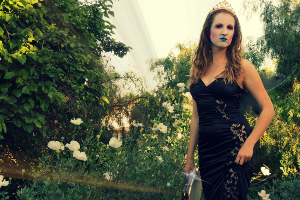 Catherine as Queen Mab in a tiara, wings, and evening dress holding her pandeiro standing in front of some white flowers.