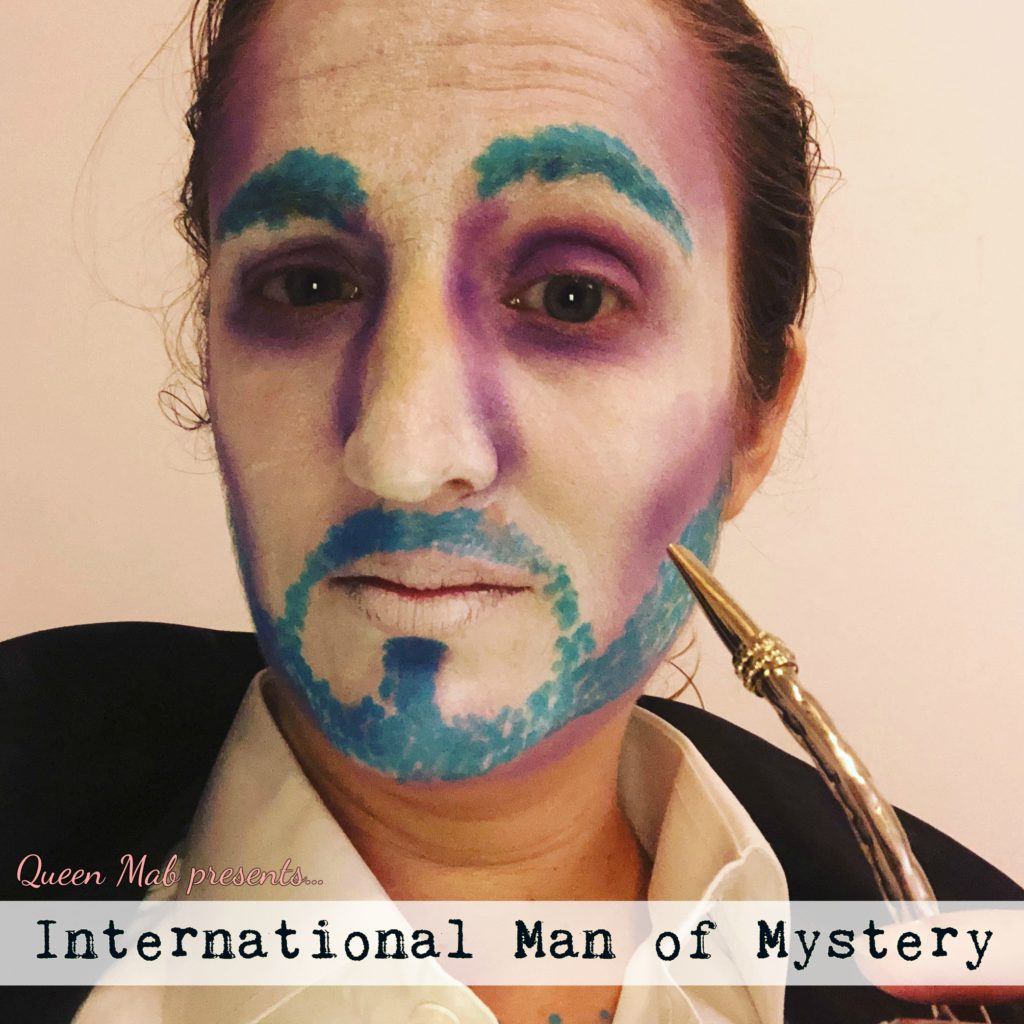 Image of Catherine with her face painted to look like a man. She has a blue beard and eyebrows and purple shadows over a white base. She has a mate straw in front of her face. Text at the bottom reads "Queen Mab presents...International Man of Mystery."
