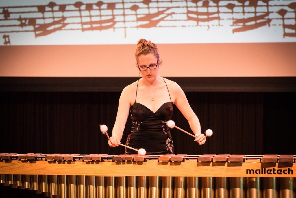 Awkward white lady in glasses and an evening dress plays a five octave marimba with four white mallets