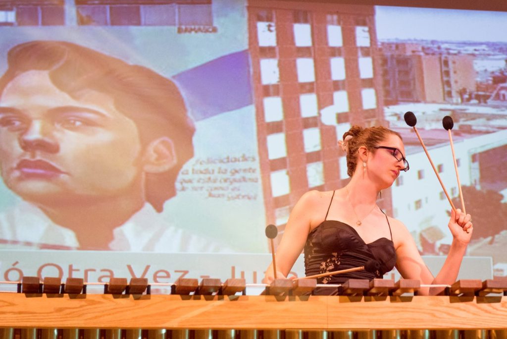 An awkward white lady with glasses and an evening dress raises her four mallets in a dramatic flourish with a mural of Juan Gabriel projected in the bakground