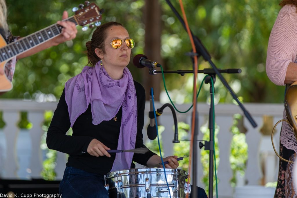 Catherine plays snare with brushes. She has on a black shirt, a purple scarf, and orange reflective sunglasses.