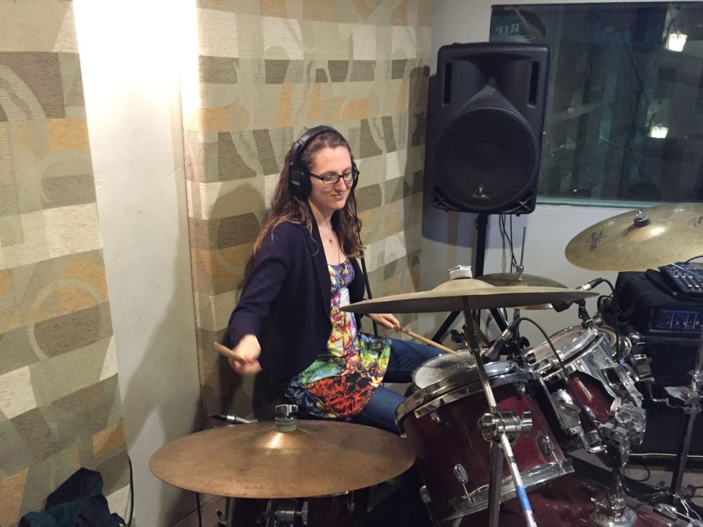 Catherine plays drum set in a recording studio booth with headphones on