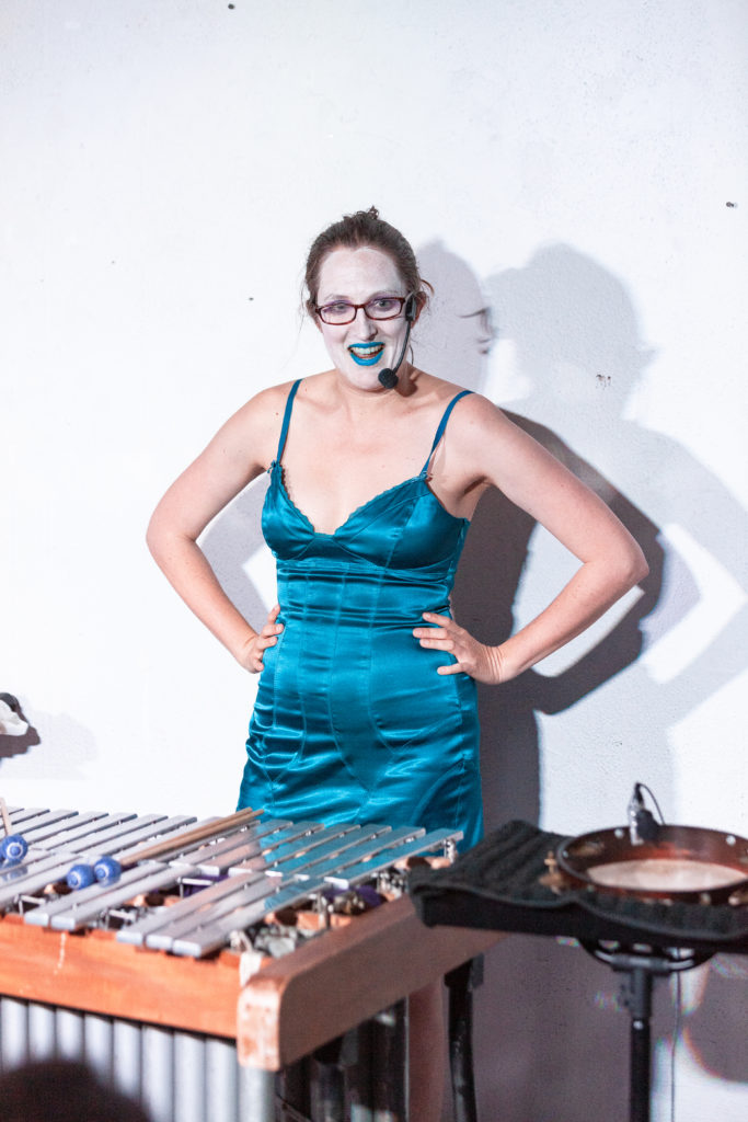 Catherine stands with her hands on her hips in front of her vibraphone and pandeiro. She is wearing a blue dress and a headset mic. She is smiling.