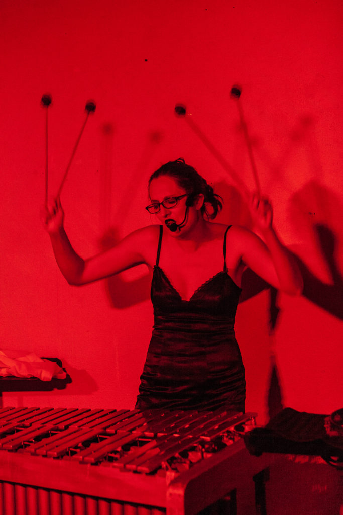Catherine plays vibraphone with four mallets. Her arms are raised. The image is lit with a red light.