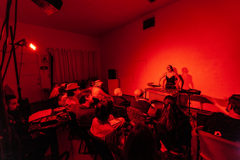 Catherine playing vibraphone and singing in front of an audience. She is lit by a red light.